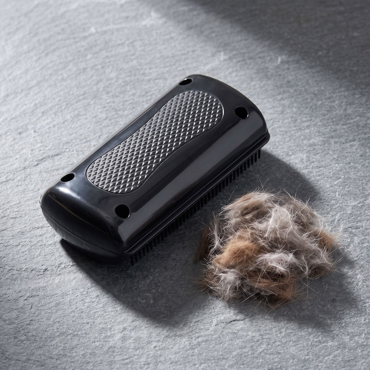 Close-up of a black pet grooming comb with dense teeth resting on a grey slate surface next to pet fur.